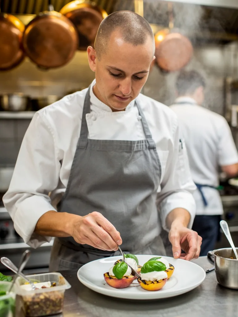 A chef with a shaved head and grey apron plating grilled peaches and burrata in a busy restaurant kitchen with copper pans and steam behind, made with FLUX.2 pro